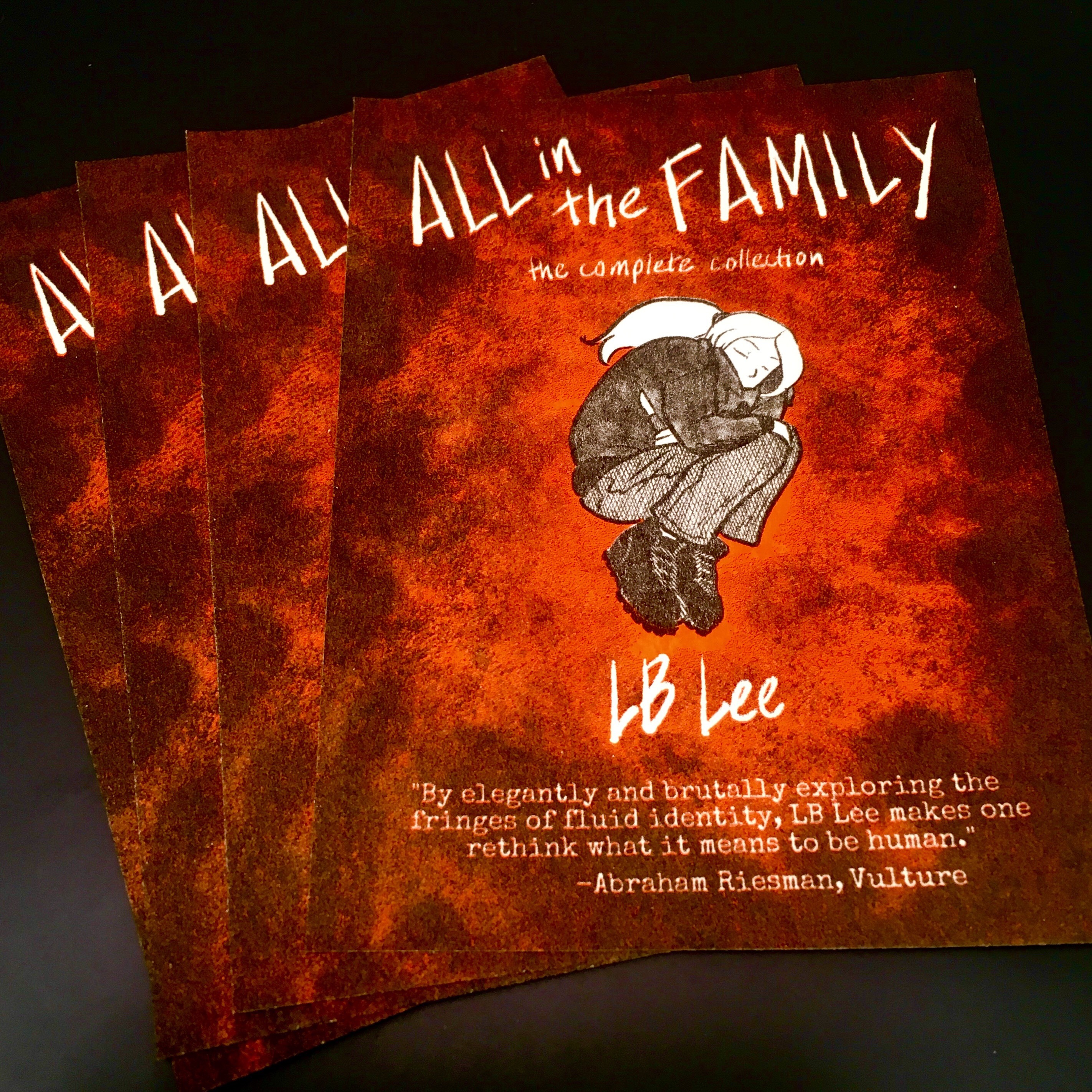 AllFam Riso Postcards A photo of a neat little stack of promotional postcards for the All in the Family omnibus, done in shades of black, white, and orange. M.D., gripping a pocket knife and dressed in combat boots, jeans, and a baggy leather jacket, floats curled in a fetal ball, the Void swirling around her in a whirlpool of smoke. With a close look, the smoke seems to be made of humanoid figures.